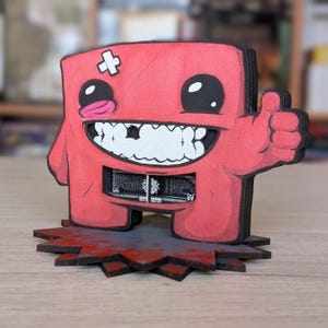 Può includere: Una figura rossa in stile cartone animato con un pollice in su. La figura ha un grande sorriso con i denti, una benda sulla testa e una lingua sporgente. Si trova su una base frastagliata grigia e rossa, con un contorno nero. È un oggetto decorativo.