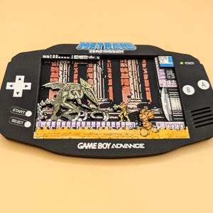 Könnte beinhalten: Ein schwarzes Game Boy Advance-Display als Schattenbox mit einer Szene aus Metroid Zero Mission. Der Bildschirmbereich zeigt ein geschichtetes 3D-Diorama mit einem geflügelten Wesen, einer kleinen Figur im gelben Anzug und einer gelben Plattform. Text Metroid Zero Mission.