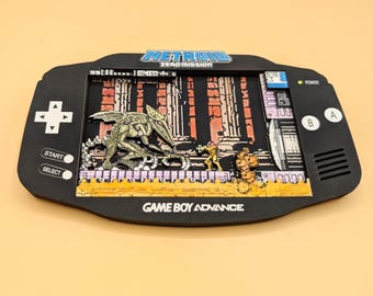 May include: A black Game Boy Advance shadow box display featuring a Metroid Zero Mission scene. The screen area shows a layered 3D diorama with a winged creature, a small character in a yellow suit, and a yellow platform. Text includes Metroid Zero Mission.