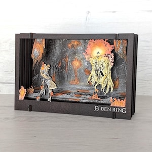 May include: A layered paper diorama depicting a scene from Elden Ring. The artwork features a fiery landscape with a character facing a large, glowing enemy. The diorama is encased in a black frame, with the game's title visible.