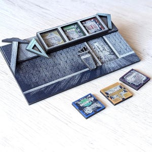 Puede incluir: Maqueta arquitectónica en miniatura detallada con una base azul oscuro y detalles plateados. El modelo presenta varias áreas enmarcadas etiquetadas como "COMMISSARY", "PATIO" y "CHAPEL". Pequeñas baldosas adicionales etiquetadas como "SECURITY", "HALLWAY" y "BOUDOIR".