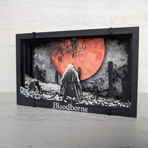Può includere: Una scatola di ombra nera con una scena di carta 3D di una figura con un lungo cappotto in piedi di fronte a una grande luna rossa. La luna ha una silhouette di albero di fronte ad essa. Il testo "Bloodborne" è stampato sul fondo della scatola.