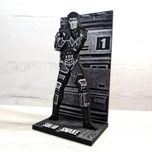 Könnte beinhalten: Eine schwarz-weiße Figur von Solid Snake aus der Metal Gear Videospielserie. Die Figur steht mit verschränkten Armen und hält eine Waffe. Die Figur steht auf einem schwarzen Sockel mit der Aufschrift "Solid Snake".