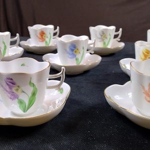 May include: A set of delicate white teacups and saucers. Each cup and saucer features a floral design with gold trim. The flowers are painted in various colors, including purple, orange, and yellow. The saucers have a unique, scalloped shape.