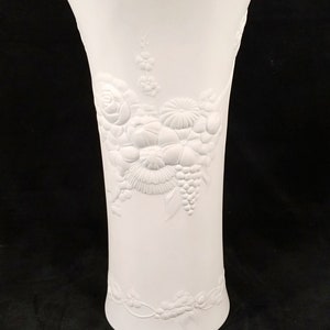 May include: White porcelain vase with an embossed floral design featuring roses, grapes, and other flowers.