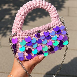 Op de afbeelding: Een lichtroze gehaakte handtas met een dik, afgerond handvat en een afneembare zilverkleurige metalen kettingriem. De voorkant van de tas is versierd met overlappende iriserende lovertjes in tinten paars, blauw en turquoise.