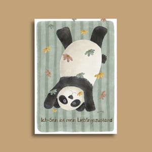 Könnte beinhalten: Eine verspielte Illustration eines Pandas, der kopfüber hängt, mit schwarz-weißem Fell und floralen Akzenten. Der Panda befindet sich vor einem hellgrünen, gestreiften Hintergrund. Der Text "Ich-Sein ist mein Lieblingszustand" ist unten.