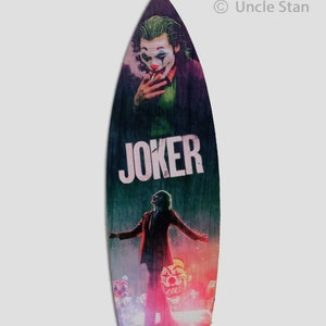 Puede incluir: Una tabla de surf de madera con un gráfico del Joker de la película "Joker". El Joker está representado con un traje verde, un chaleco morado y maquillaje blanco. La tabla de surf tiene la palabra "JOKER" impresa en letras blancas en la parte delantera. El Joker también está representado con un traje rojo, una camisa blanca y una corbata negra, de pie sobre un coche de policía con un cartel que dice "KILL THE" delante de él.