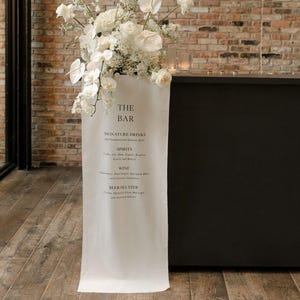 Könnte beinhalten: Ein weißes Stoffschild mit der Aufschrift "THE BAR" und einer Getränkekarte. Das Schild ist mit weißen Blumen geschmückt und steht vor einer Backsteinmauer. Die Bar ist schwarz.
