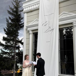May include: A long white fabric banner hangs from a building balcony, featuring the printed text 'WELCOME TO OUR FOREVER', the names 'Ed & Biky', and the date '06.10.2024'. The banner extends down to the grass where two people stand reading papers.
