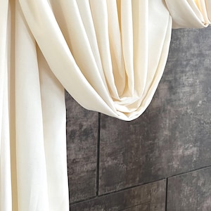 May include: Off-white fabric draped over a wooden rail, forming gentle folds and shadows. The fabric hangs against a textured, dark grey wall, suggesting a home decor or textile product.