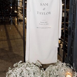 May include: A white wedding sign with black text announcing the wedding of Sam and Taylor in Duluth, Minnesota on October 11, 2024. The sign is displayed on a black stand with white baby's breath flowers on either side.