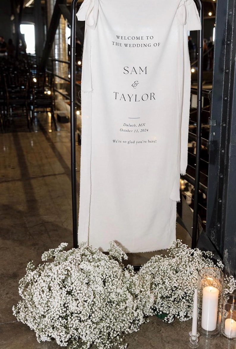 May include: A white wedding sign with black text announcing the wedding of Sam and Taylor in Duluth, Minnesota on October 11, 2024. The sign is displayed on a black stand with white baby's breath flowers on either side.