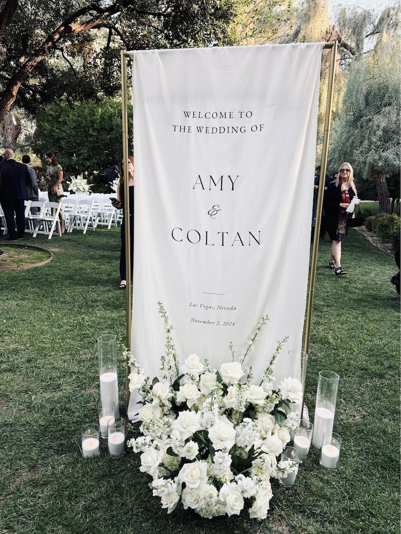 May include: A white wedding sign with gold trim, featuring the text "Welcome to the wedding of Amy & Coltan", "Las Vegas, Nevada", and "November 2, 2024". The sign is decorated with white flowers and candles.
