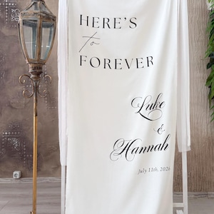 May include: A white fabric wedding sign hanging on a white metal frame. The text reads: HERE'S to FOREVER, Luke & Hannah, 11th July 2026. The sign features black lettering in a mix of serif and script fonts, with raw edges at the bottom of the fabric.