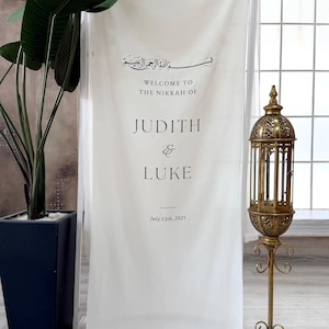 May include: A white fabric banner with black text that reads "WELCOME TO THE NIKKAH OF JUDITH & LUKE July 11th, 2025." The banner is displayed on a clear frame. A decorative gold lantern and potted plant are also visible.