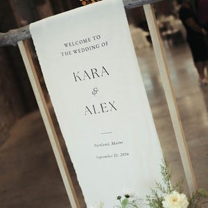 May include: A white sign with black text announcing the wedding of Kara and Alex in Portland, Maine on September 21, 2024. The sign is attached to a wooden frame with white flowers in the foreground.