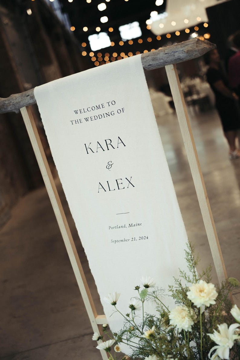 May include: A white sign with black text announcing the wedding of Kara and Alex in Portland, Maine on September 21, 2024. The sign is attached to a wooden frame with white flowers in the foreground.