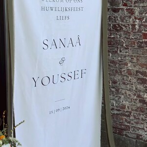 May include: A white sign with black text announcing a wedding. The text reads "Marhaba bikom" (Arabic for "Welcome"), "Welkom op ons huwelijksfeest" (Dutch for "Welcome to our wedding"), "Liefs" (Dutch for "Love"), "Sanaa & Youssef", and "15/09/2024".