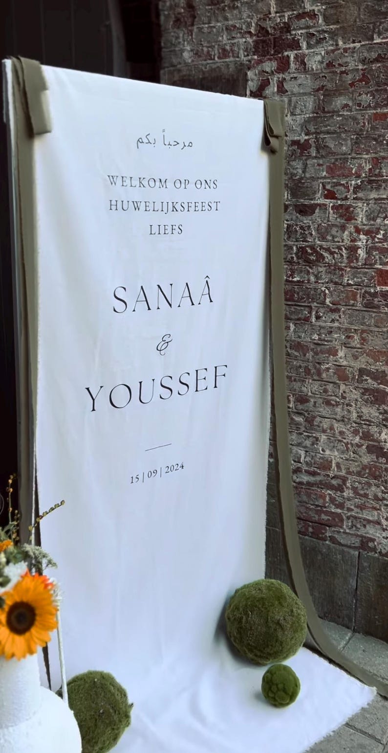 May include: A white sign with black text announcing a wedding. The text reads "Marhaba bikom" (Arabic for "Welcome"), "Welkom op ons huwelijksfeest" (Dutch for "Welcome to our wedding"), "Liefs" (Dutch for "Love"), "Sanaa & Youssef", and "15/09/2024".
