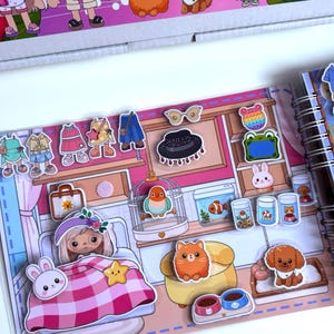May include: A paper doll activity book featuring a bedroom scene with various removable stickers including clothing, accessories, a bird in a cage, a fish tank, a cat, a dog, and a character in a bed with a pink plaid blanket and a yellow star.