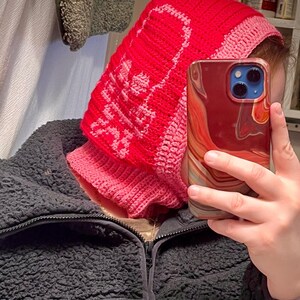Op de afbeelding: Een roze en rode gebreide sjaal met capuchon met een schedelontwerp. De sjaal wordt gedragen door een persoon die een rood, wit en blauw gemarmerde telefoon vasthoudt.