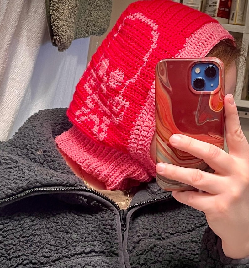 Op de afbeelding: Een roze en rode gebreide sjaal met capuchon met een schedelontwerp. De sjaal wordt gedragen door een persoon die een rood, wit en blauw gemarmerde telefoon vasthoudt.