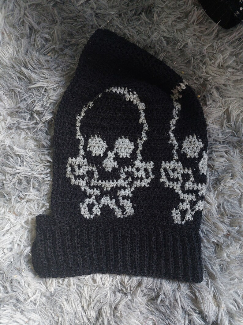 Op de afbeelding: Zwarte gebreide beanie met een wit schedelontwerp. De schedel wordt twee keer op de beanie herhaald.