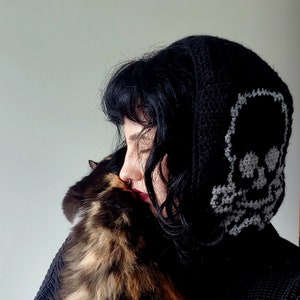 Op de afbeelding: Een persoon draagt een zwarte hoodie met een wit schedelontwerp op de capuchon, terwijl ze een driekleurige kat vasthoudt.