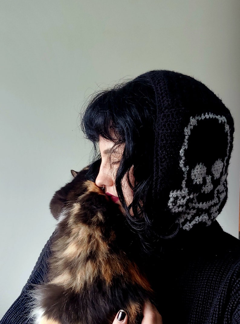 Op de afbeelding: Een persoon draagt een zwarte hoodie met een wit schedelontwerp op de capuchon, terwijl ze een driekleurige kat vasthoudt.