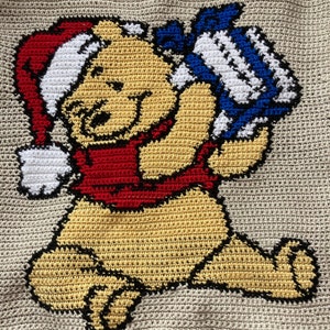 May include: A crocheted Winnie the Pooh character wearing a red and white Santa hat and holding a blue and white gift. The character is sitting on a beige background.