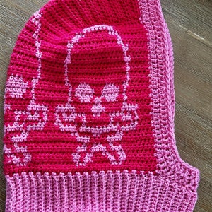 Op de afbeelding: Een roze gebreide muts met een schedelontwerp. De muts heeft een roze en wit schedelpatroon aan de voorkant en een effen roze rand.