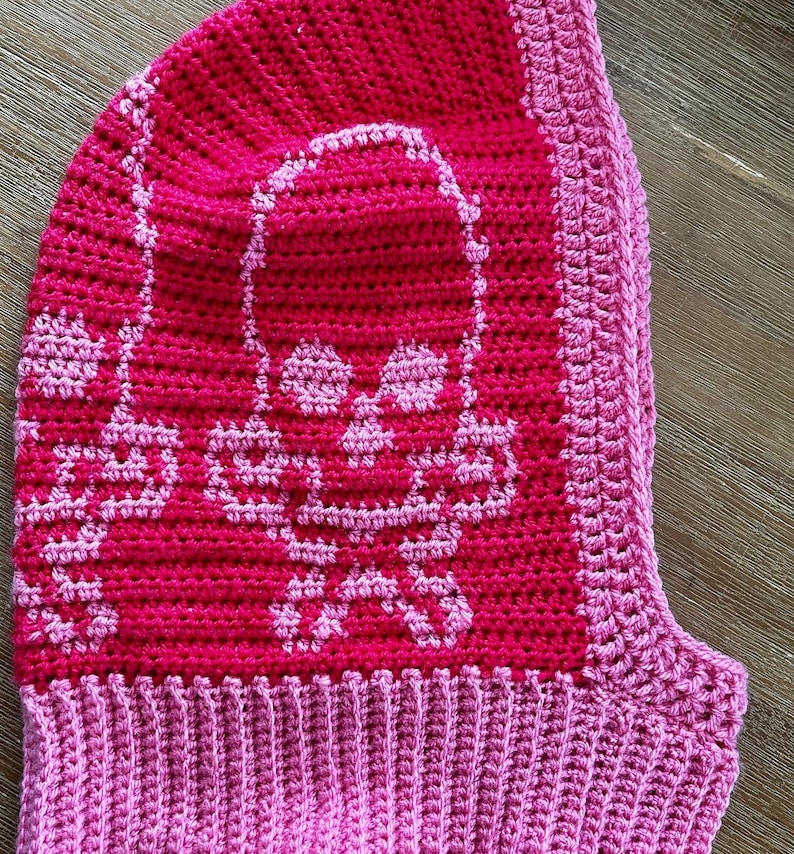 Op de afbeelding: Een roze gebreide muts met een schedelontwerp. De muts heeft een roze en wit schedelpatroon aan de voorkant en een effen roze rand.