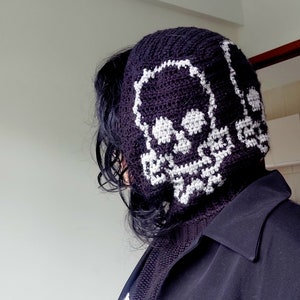 Op de afbeelding: Een zwarte gehaakte beanie met een wit schedelontwerp. De beanie wordt gedragen door een persoon met lang zwart haar.