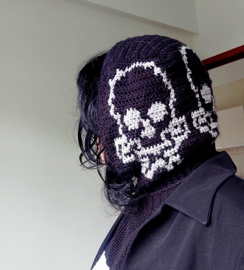 Op de afbeelding: Een zwarte gehaakte beanie met een wit schedelontwerp. De beanie wordt gedragen door een persoon met lang zwart haar.