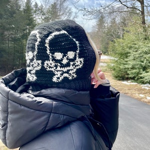 Op de afbeelding: Zwarte gehaakte beanie met een wit schedelontwerp. De beanie wordt gedragen door een persoon in een zwarte pufferjas.