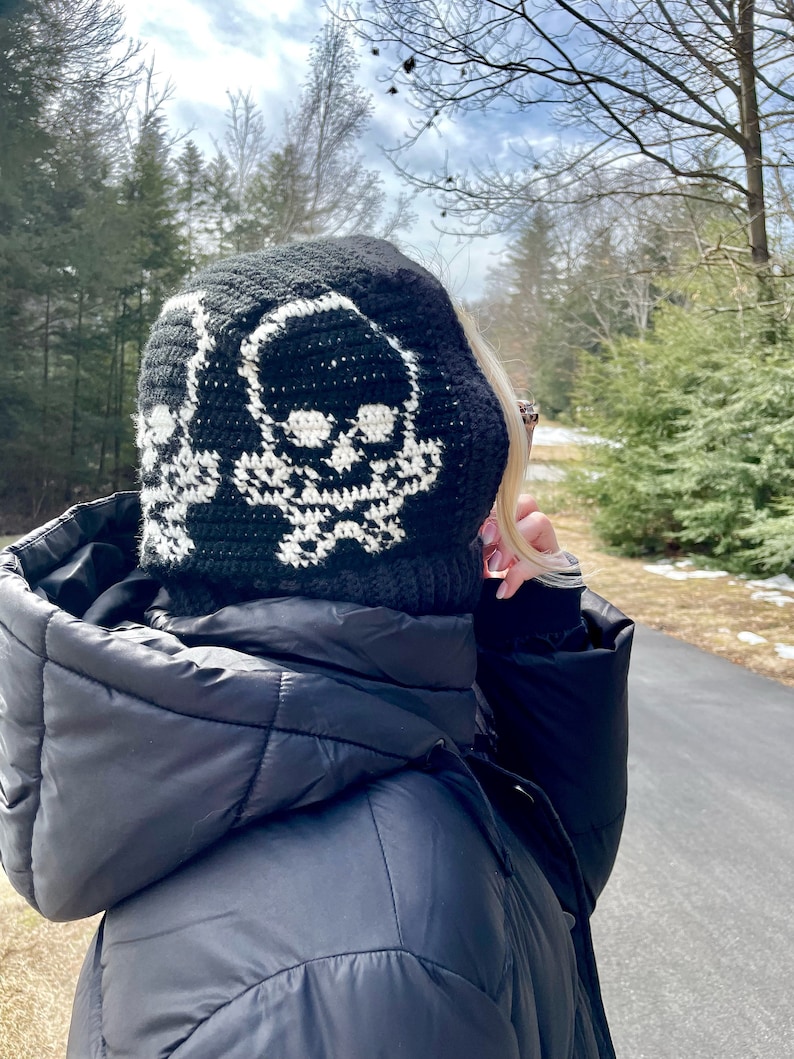 Op de afbeelding: Zwarte gehaakte beanie met een wit schedelontwerp. De beanie wordt gedragen door een persoon in een zwarte pufferjas.