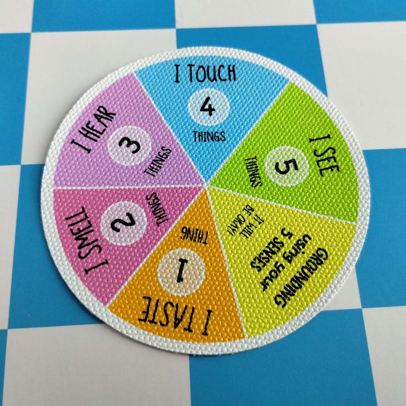 May include: A circular grounding tool featuring a colorful pie chart design. The segments are labeled with sensory prompts: I TASTE 1 THING, I SMELL 2 THINGS, I HEAR 3 THINGS, I TOUCH 4 THINGS, and I SEE 5 THINGS, with the text IT WILL BE OKAY.