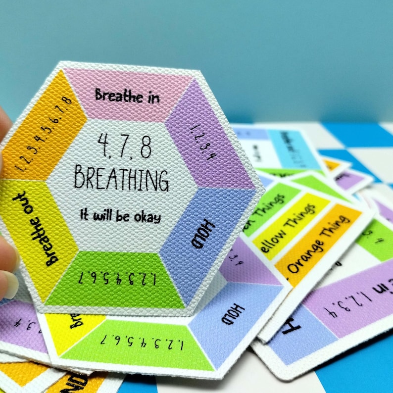 May include: A hexagonal card with a textured white surface featuring the text 4, 7, 8 BREATHING and It will be okay. The card is divided into colored sections including pink, purple, blue, green, yellow, and orange with printed numbers and instructions.
