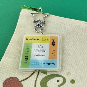 K&ouml;nnte beinhalten: Ein quadratischer Acryl-Schl&uuml;sselanh&auml;nger mit einer Anleitung zur Box-Atmung und dem Text Breathe in 1, 2, 3, 4, HOLD 1, 2, 3, 4, Breathe out 1, 2, 3, 4, BOX BREATHING und It will be okay, befestigt an einem sternf&ouml;rmigen Metallverschluss auf einer Stofftasche.