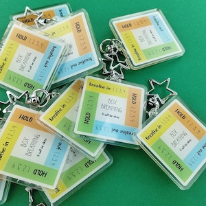 May include: A collection of square acrylic keychains featuring a box breathing guide. Each keychain has a star-shaped clasp and displays the text: "BOX BREATHING, It will be okay," surrounded by color-coded sections labeled "Breathe in 1, 2, 3, 4," "HOLD 1, 2, 3, 4," "Breathe out 1, 2, 3, 4," and "HOLD 1, 2, 3, 4."