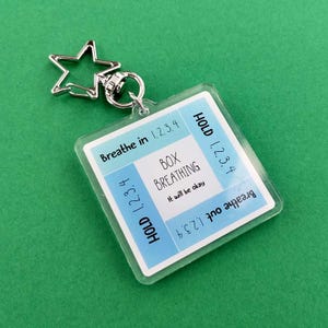 May include: A clear square keychain with a silver star-shaped charm. The keychain has a blue and white design with the words "Breathe in 1.2.3.4", "Hold 1.2.3.4", "Box Breathing", "It will be okay", and "Breathe out 1.2.3.4".