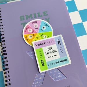 May include: A purple spiral-bound notebook with a circular sticker featuring a 5-4-3-2-1 grounding technique wheel and a square sticker with a box breathing guide. The square sticker includes the text Breathe in, Hold, Breathe out, and Box Breathing.