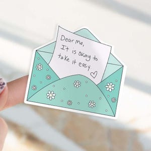 May include: A sticker in the shape of a teal envelope with white daisy accents. A white note inside reads "Dear me, It is okay to take it easy." A small heart is drawn at the end of the message.