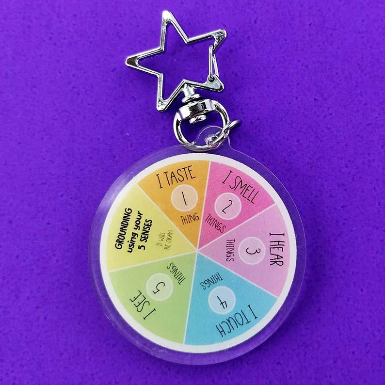 K&ouml;nnte beinhalten: Ein runder Acryl-Schl&uuml;sselanh&auml;nger mit einem sternf&ouml;rmigen silbernen Metallverschluss. Das Design ist ein buntes Rad, das in f&uuml;nf Segmente unterteilt ist, beschriftet mit den 5 Sinnen: I TASTE 1 THING, I SMELL 2 THINGS, I HEAR 3 THINGS, I TOUCH 4 THINGS, I SEE 5 THINGS.