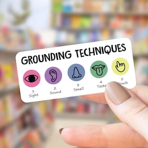 May include: A rectangular white card featuring the black text GROUNDING TECHNIQUES. Below are five colored circles with black icons: a pink eye for 1 Sight, a purple ear for 2 Sound, a blue nose for 3 Smell, a green mouth for 4 Taste, and a yellow hand for 5 Touch.