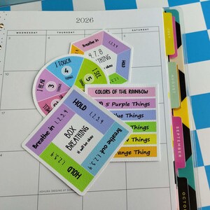May include: Three square and circular stickers with grounding techniques and mindfulness exercises. The stickers feature text including Box Breathing, 4-7-8 Breathing, and Colors of the Rainbow, with colorful sections in pink, blue, green, yellow, and orange.