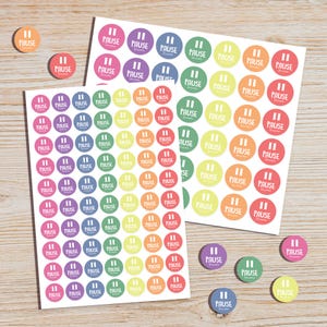 May include: Colorful round stickers with the word "PAUSE" and a pause symbol, in various colors like pink, purple, green, yellow, orange, and blue. The stickers are on a white sheet and scattered on a wooden surface.