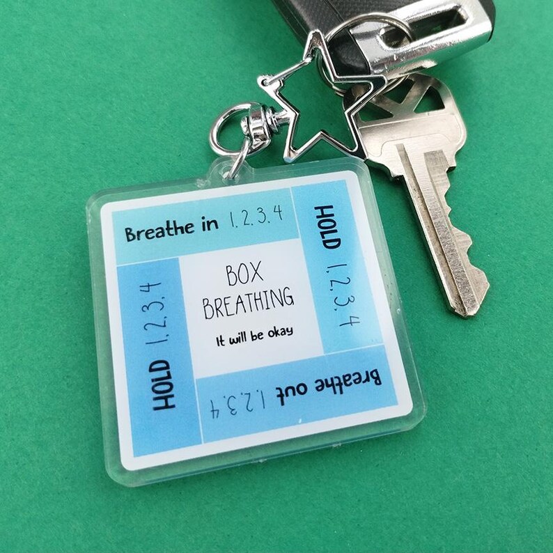 May include: A clear, square keychain with a blue and white design. The keychain features the words "Breathe in 1.2.3.4", "Hold 1.2.3.4", "Box Breathing", "It will be okay", and "Breathe out 1.2.3.4". A silver star-shaped charm and keys are attached.