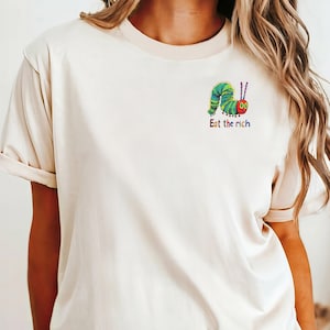 Op de afbeelding: Een wit T-shirt met een kleurrijke rups afbeelding en de tekst "Eat the rich" gedrukt op de voorkant.